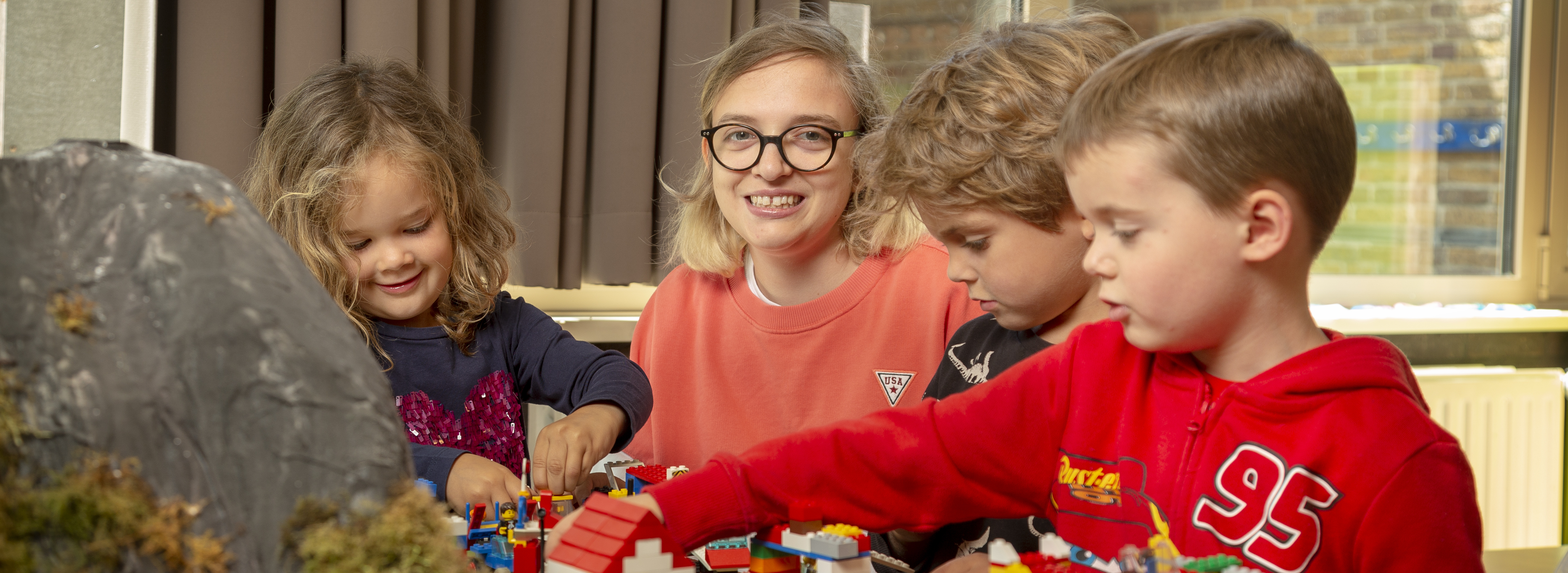 Persoon met een bril op lacht naar de camera en zit aan tafel vol met lego te spelen met 3 kinderen.